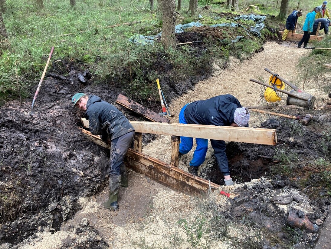 Zwei Mitarbeiter beim Aufbau einer Holzbohlenwand im Graben-