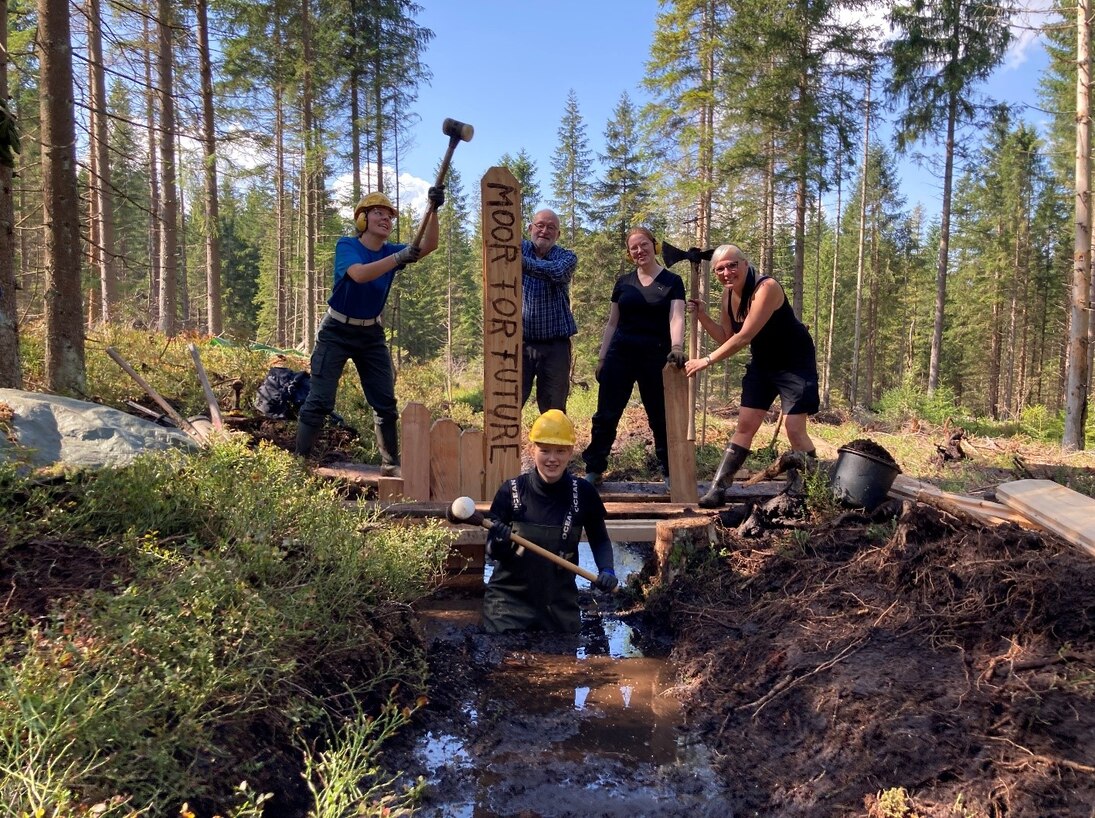 Fünf Mitarbeiter posieren mit ihren Arbeitsgeräten und einem Schild mit der Aufschrift "Moor for future" in einem Wassergraben.